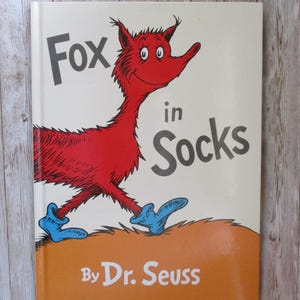 May include: A hardcover book titled "Fox in Socks" by Dr. Seuss. The cover features a red fox-like creature wearing blue socks, with the title in large, bold letters. The background is a light cream color with an orange band at the bottom.