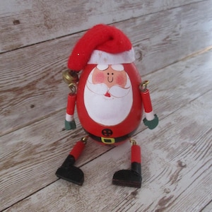 May include: A wooden Santa Claus figurine with a red body, hat, and articulated limbs. The Santa has a white beard, a smiling face, and a black belt. The figurine is wearing black boots and green gloves. A small gold bell hangs from the arm.
