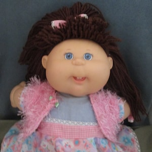 May include: A Cabbage Patch Kid doll with brown yarn hair, wearing a pink fuzzy jacket and a floral dress with a pink and white gingham trim.
