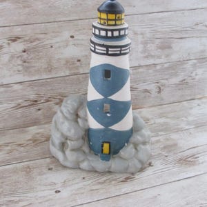 May include: A ceramic lighthouse figurine with a black top and yellow light. The lighthouse has a white and blue body, and sits on a gray rock base. The lighthouse is a decorative nautical item.