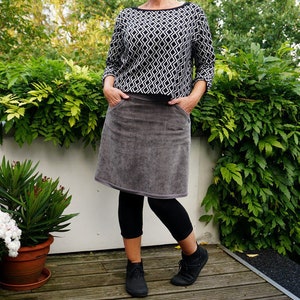 May include: A woman wearing a gray corduroy skirt with black leggings and black ankle boots. She is wearing a black and white geometric patterned sweater.