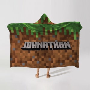 May include: A hooded blanket featuring a pixelated design reminiscent of a video game landscape. The blanket is primarily brown, with green grass at the top and the name "JONATHAN" in gray block letters. The blanket is being worn.