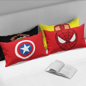 May include: Four rectangular decorative pillows with superhero designs on a white bed. The pillows feature Captain America's shield, Spider-Man's mask, and other comic book characters. A book is open on the bed.