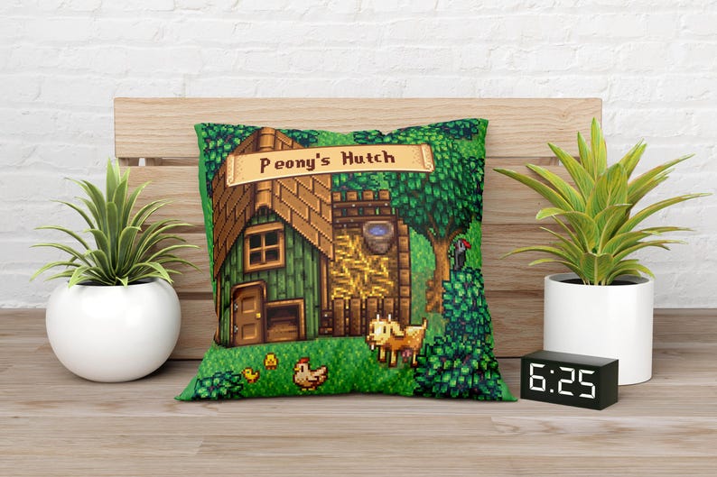 Puede incluir: Un coj&iacute;n decorativo verde con una ilustraci&oacute;n pixelada de un edificio con la etiqueta "Peony's Hutch". El dise&ntilde;o incluye un tejado marr&oacute;n, una puerta de madera y una cabra. Tambi&eacute;n se ven dos plantas en maceta y un reloj digital.
