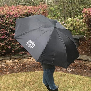 May include: A black umbrella with a white monogram on the top. The umbrella is being held by a person wearing black rain boots with a white monogram.