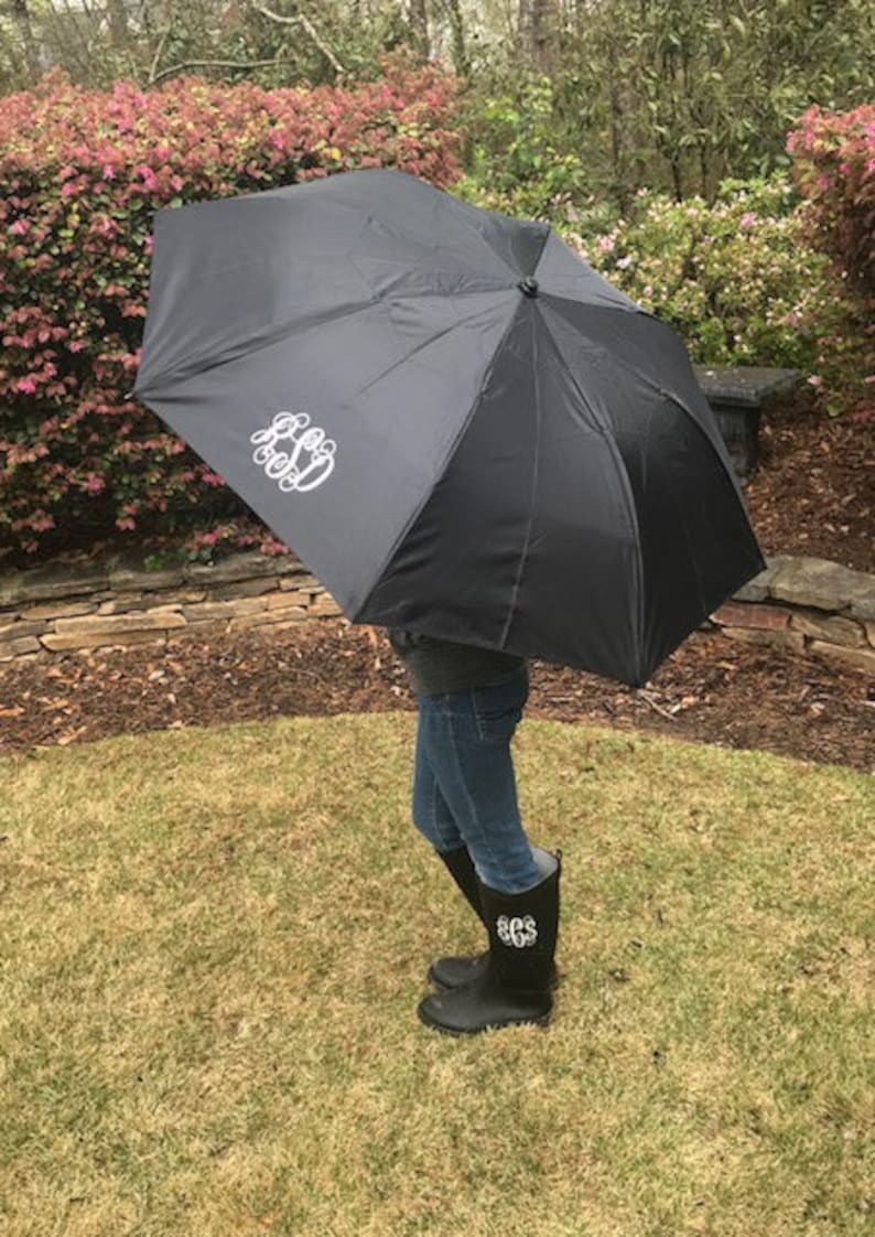 May include: A black umbrella with a white monogram on the top. The umbrella is being held by a person wearing black rain boots with a white monogram.