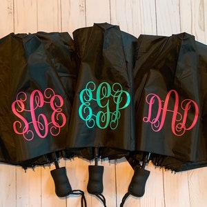 May include: Three black umbrellas with personalized monograms. The monograms are in pink, teal, and turquoise. The umbrellas are folded and have black handles.