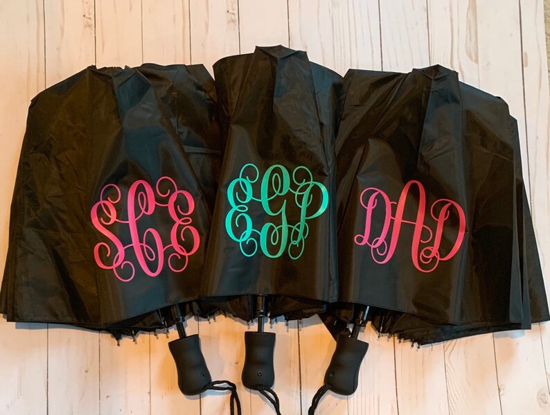 May include: Three black umbrellas with personalized monograms. The monograms are in pink, teal, and turquoise. The umbrellas are folded and have black handles.