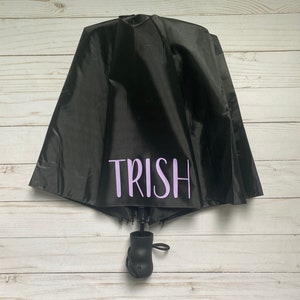 May include: A black umbrella with a purple inscription that reads "TRISH".