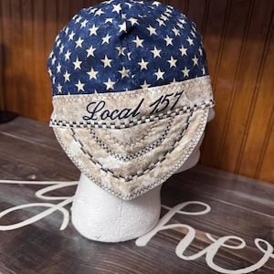 May include: A fabric welding cap featuring a dark blue crown with a repeating pattern of cream-colored stars. The lower band is a textured beige fabric with the embroidered text Local 157 in dark blue, accented by decorative zigzag stitching.