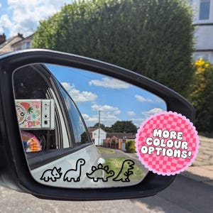 May include: Car side mirror with a black outline of four dinosaurs. A pink sticker with the text "MORE COLOUR OPTIONS!" is on the right. The mirror reflects a blue sky and houses.