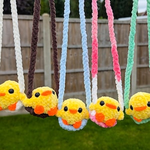 May include: Five crocheted yellow chicks with orange beaks and black eyes, each attached to a long, twisted yarn strap in white, brown, light blue, pink, and mint green, hanging in a row against a wooden fence background.