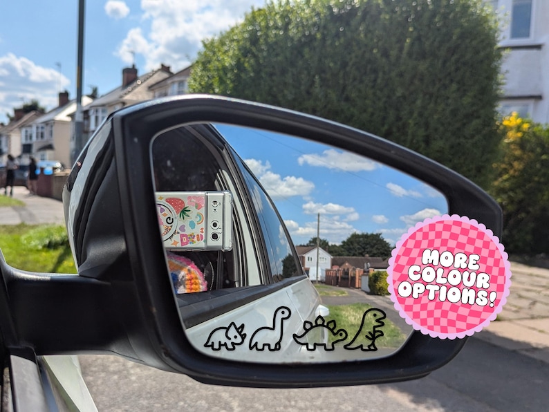 May include: Car side mirror with a black outline of four dinosaurs. A pink sticker with the text "MORE COLOUR OPTIONS!" is on the right. The mirror reflects a blue sky and houses.