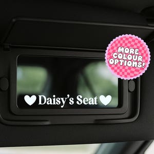 May include: Black car visor with a mirror, featuring a white decal that reads "Daisy's Seat" between two white hearts. A pink circle with the text "MORE COLOR OPTIONS!" is attached to the visor.