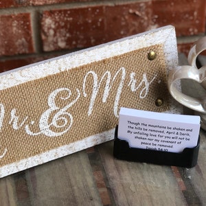 May include: A white wooden sign with burlap fabric and the words "Mr. & Mrs." in white paint. The sign is sitting on a wooden surface next to a white ceramic heart sculpture and a card with a Bible verse about love.