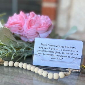 May include: A white card with a blue background and black text. The text reads "Peace I leave with you Elizabeth; My peace I give you. I do not give to you as the world gives. Do not let your heart be troubled and do not be afraid John 14:27". The card is sitting in a clear plastic holder on a wooden surface.