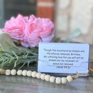 May include: A white card with a bible verse printed on it. The verse reads "Though the mountains be shaken and the hills be removed, Brittany, My unfailing love for you will not be shaken nor my covenant of peace be removed. Isaiah 54:10". The card is sitting on a table with a wooden bead garland in front of it.