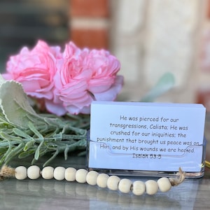 May include: A white card with a bible verse printed on it. The verse is Isaiah 53:5. The card is sitting on a clear acrylic stand. The stand is surrounded by white beads and pink flowers.