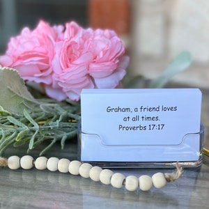 May include: A white card with the text "Graham, a friend loves at all times. Proverbs 17:17" in black font. The card is in a clear plastic holder on a wooden surface with a string of wooden beads.