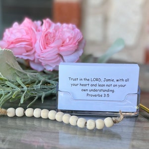 May include: A clear acrylic card holder with a stack of white cards. The cards have text that reads "Trust in the LORD, Jamie, with all your heart and lean not on your own understanding. Proverbs 3:5". A wooden bead garland and pink flowers are in the background.