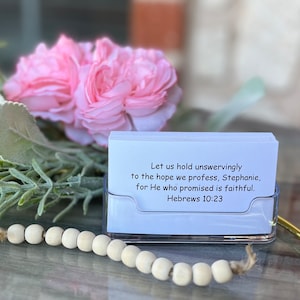 May include: White card with a bible verse printed on it. The verse reads "Let us hold unswervingly to the hope we profess, Stephanie, for He who promised is faithful. Hebrews 10:23". The card is sitting in a clear plastic holder on a table with a string of wooden beads.