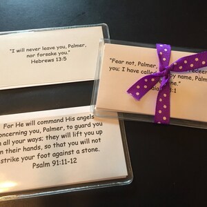 May include: Clear plastic card holders with printed text. One card holder has a purple ribbon tied around it. The text includes Bible verses from Hebrews 13:5, Isaiah 43:1, and Psalm 91:11-12. The cards are on a dark surface.