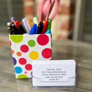 May include: A desk organizer with colorful pens, markers, and pink scissors. A small clear card holder displays a stack of white cards with the text "Fear not, Julie, for I have redeemed you; I have called you by name. Julie, you are mine. Isaiah 43:1."