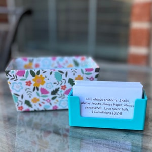 May include: A teal blue card holder with a stack of white cards inside. The top card has a quote about love from 1 Corinthians 13:7-8. The card holder is sitting on a glass table with a floral patterned box behind it.