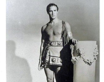 May include: Black and white photograph of a man in a Roman-style costume. He wears a short skirt, sandals, and armbands, and leans against a decorative column. The image includes a signature in red ink.