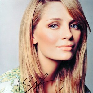 May include: A close-up portrait of a woman with long, straight, light blonde hair. She is wearing a floral blouse and has a natural makeup look. Her eyes are blue and she is smiling slightly. The image is signed with a black signature.