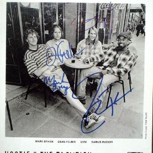 Könnte beinhalten: Ein Schwarzweißfoto von vier Männern, die an einem Tisch im Freien sitzen. Die Männer lächeln alle und schauen in die Kamera. Das Foto ist von jedem der Männer signiert, die Mitglieder der Band Hootie & The Blowfish sind. Der Name der Band ist unter dem Foto zusammen mit dem Logo von Atlantic Records gedruckt.