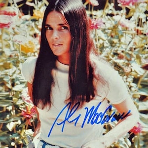 以下が含まれることがあります： 長い黒髪の女性が白いTシャツとブルージーンズを着ているヴィンテージ写真。青いインクでサインされており、白いベルトと花の背景があります。