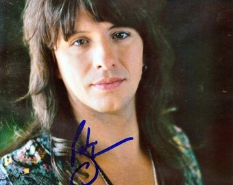 May include: A close-up portrait of a man with long brown hair, wearing a blue and green paisley patterned shirt with a black beaded necklace. The man is looking at the camera and has a slight smile on his face. The image is signed in blue ink.