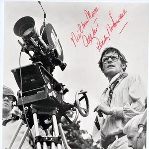 May include: A black and white photo of a man standing next to a film camera on a tripod. The man is wearing a light-colored shirt and glasses. He is holding a microphone in his right hand. The camera is pointed towards the sky. The man is signing his name "Nic' Plowman" and "All the best" in red ink.