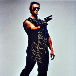 Puede incluir: Una fotografía en blanco y negro de un hombre con gafas de sol y una camiseta negra sin mangas sosteniendo un arma. El hombre mira a la cámara con una expresión seria. La fotografía está firmada "Arnold Schwarzenegger".