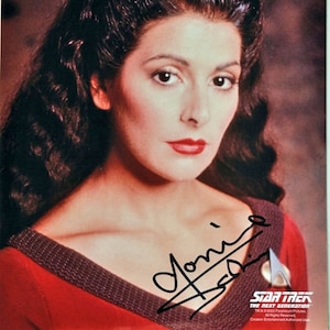 May include: A close-up portrait of a woman with long dark hair styled in a bun with a blue and red hair accessory. She is wearing a red and black uniform with a Starfleet insignia. The woman is smiling and has red lipstick. The image is signed "Joni" in black ink.