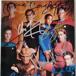 May include: A color photograph featuring a group of people, likely actors, posing together. The image is signed by multiple people. The subjects are dressed in various costumes, suggesting a science fiction theme. The background is a warm, golden hue.