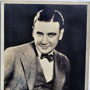 May include: A black and white photograph of a man wearing a suit and bow tie. He is smiling and looking at the camera. The photo is signed "Sincerely, Richard Dix".