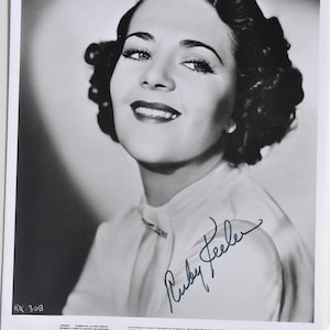 May include: Black and white photograph of Ruby Keeler, a woman with dark, curly hair and a radiant smile. She wears a light-colored blouse with a brooch. The image is signed and includes text at the bottom.
