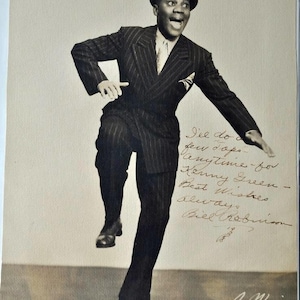 May include: Black and white photograph of a man in a pinstripe suit and bowler hat, mid-dance. The image includes handwritten text and a signature. The man is smiling with one leg raised.