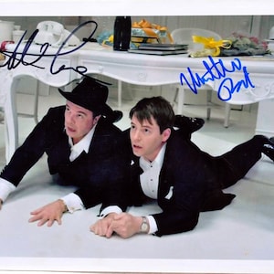 May include: A black and white photo of two men in suits, lying on the floor under a white table. The men are looking at the camera and smiling. The photo is signed by both men, "Matthew Perry" and "Wattle Burt".