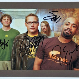 Könnte beinhalten: Gerahmtes Foto mit vier Männern, vermutlich eine Band, die vor einer Holzwand posieren. Jedes Mitglied hat das Foto signiert. Der Mann rechts trägt ein braunes T-Shirt mit dem Text "THE SILOS".