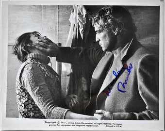 May include: Black and white photograph of a man and woman in a scene from a movie. The man is holding the woman's face with his hand. The man's signature, "Marlon Brando", is written in blue ink on the image.