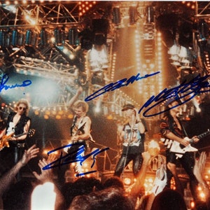 May include: A photograph of a rock band performing on stage, with bright stage lights and a crowd in the foreground. The band members are wearing stage outfits and playing instruments. The photo has blue signatures on it.