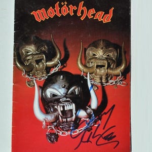 May include: A vintage album cover featuring the band Motörhead. The cover has a red background with three menacing, horned skull figures. The band's name is written in a stylized, red font at the top. The cover also includes a signature.