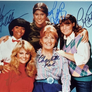 May include: A colour photograph of six people, possibly a television cast, posing together. Each person is smiling and wearing different clothing. The photo has several signatures in blue ink across the top.
