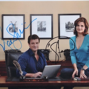May include: A signed photograph of two actors, a man wearing a blue shirt and a woman wearing a blue shirt. The man is sitting at a desk with a laptop and a book. The woman is standing behind the desk with a book in her hand. The photo is signed by both actors.
