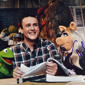 May include: A man in a plaid shirt sits at a table with a green Muppet and a pink Muppet. The man is holding a script and the pink Muppet is holding a pen. The pink Muppet has blonde hair and is wearing a black jacket. The green Muppet is wearing a red mouth and has a green body.