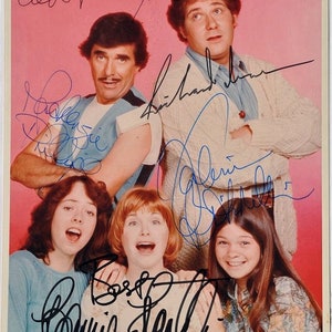 May include: A signed photograph of the cast of the television show 'The Brady Bunch'. The cast members are shown in a group pose, with the three girls in the front row and the three men in the back row. The photo is signed by all six cast members: Robert Reed, Ann B. Davis, Florence Henderson, Christopher Knight, Barry Williams, and Mike Lookinland.