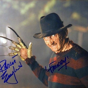 Può includere: Fotografia a colori di un uomo con un cappello fedora e un maglione a righe, con un guanto con artigli di metallo sulla mano destra. L'uomo sorride. L'immagine include le firme "Robert Englund" e "Freddy".